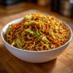 Close-up of a white bowl filled with savory, saucy fried noodles mixed with shredded carrots and cabbage.