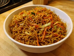 A close-up of a white bowl filled with glossy, savory fried noodles mixed with shredded carrots and cabbage.