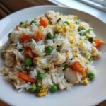 A close-up of a white plate filled with homemade chicken fried rice featuring white rice, peas, carrots, and scrambled egg pieces.