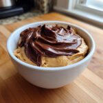Close-up of a white bowl filled with creamy peanut butter dip topped with a thick swirl of melted chocolate.