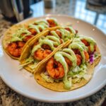 Three appetizing shrimp taco shells filled with seasoned shrimp, slaw, and drizzled generously with green avocado crema.