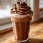A tall glass of rich, frothy whipped hot chocolate topped with a swirl of chocolate whipped cream.