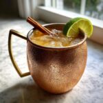 A frosty copper mug filled with an apple cider moscow mule, garnished with a lime wedge and cinnamon stick.