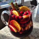 A clear glass pitcher filled with vibrant Christmas sangria, garnished with oranges, cranberries, apples, cinnamon sticks, and rosemary.