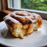 A close-up of a gooey cinnamon roll apple pie slice topped with white icing, sitting on a white plate.