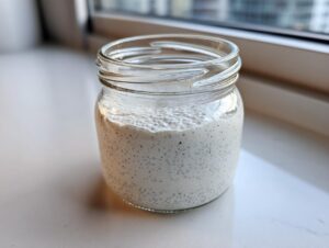 A glass jar filled with creamy, white coconut chia pudding, set on a white surface near a window.