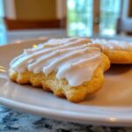 A close-up of a golden sugar cookie topped with thick, white, slightly drippy cookie icing on a white plate.