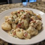 A close-up of creamy crack chicken gnocchi served on a white plate, topped with bacon bits and parsley.