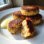 Four golden-brown, crispy salmon cakes stacked on a white plate, one cut open to show the tender interior, served with a side of yellow sauce.