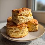Four tall, flaky, golden-brown scones stacked on a light ceramic plate, showing off their layers.