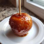 A single, glossy caramel apples coated in rich, dripping caramel sauce, served on a white plate.