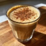 A close-up of a glass filled with creamy, frothy coquito, topped generously with ground cinnamon.