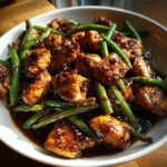 Close-up of bite-sized pieces of glazed chicken and bright green beans tossed in a dark, savory sauce, ready to eat.