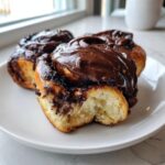 Three soft, freshly baked chocolate cinnamon rolls heavily topped with glossy chocolate frosting, one showing a bite taken out.