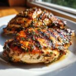 Close-up of two grilled chicken breasts coated in a dark, herb-flecked chicken marinade, glistening in sunlight.