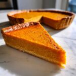 A close-up slice of vibrant orange sweet potato pie resting on a white marble surface, with the rest of the pie blurred in the background.