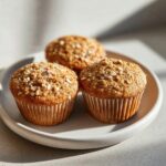 Three golden brown oatmeal muffins topped with rolled oats, sitting on a light ceramic plate.