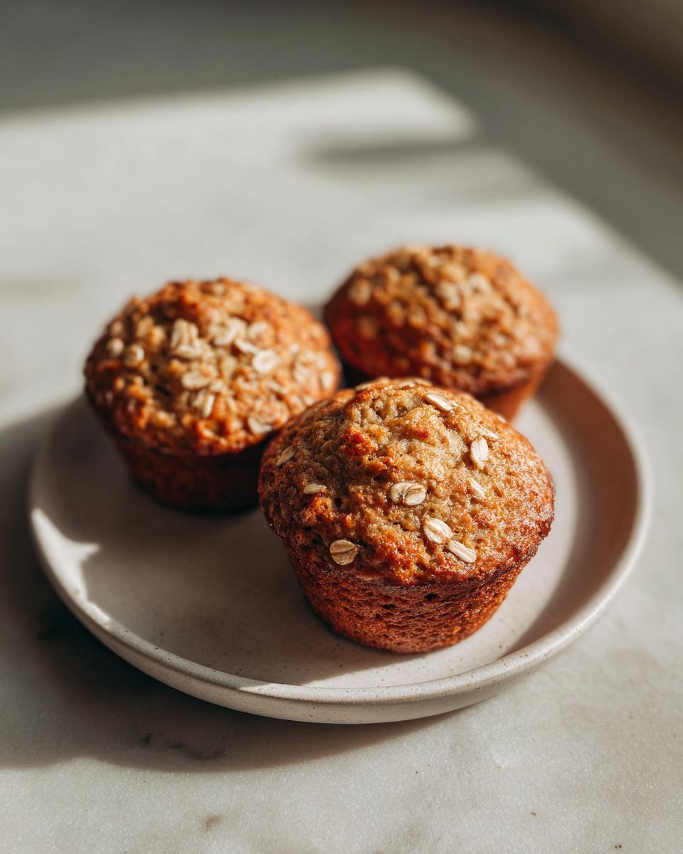 Three golden brown oatmeal muffins topped with rolled oats, sitting on a light ceramic plate.