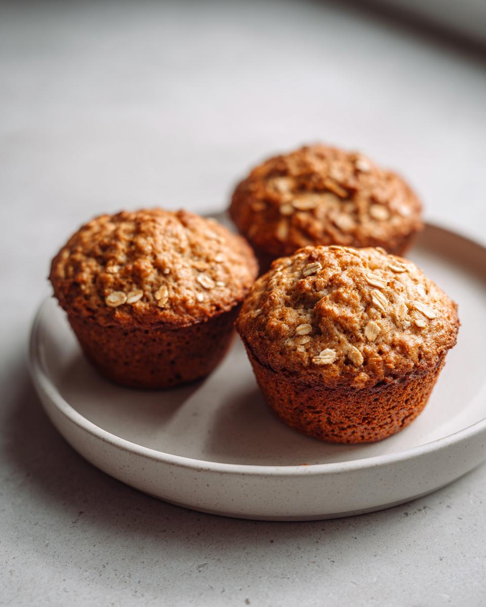Three freshly baked, moist oatmeal muffins topped with rolled oats sitting on a light-colored speckled plate.
