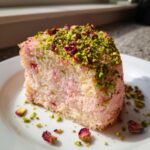 A square slice of moist, pink rose cake topped generously with chopped pistachios and dried rose petals.