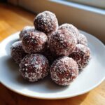 A close-up stack of rich, dark no-bake bourbon balls heavily dusted with white powdered sugar on a white plate.