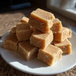 A pile of freshly cut, light brown penuche fudge squares stacked on a white plate.