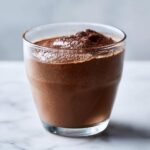 A close-up of rich, airy chocolate mousse served in a clear glass, topped with a dusting of cocoa powder.