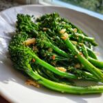 Close-up of bright green roasted broccolini tossed with crispy garlic pieces on a white plate.