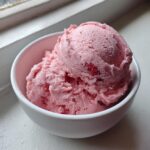 Two scoops of bright pink strawberry ice cream with visible fruit pieces served in a small white bowl.