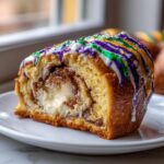 Close-up of a slice of king cake showing cream cheese filling, cinnamon swirl, and purple, green, and gold icing.