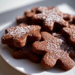 A close-up of several rich brown soft gingerbread cookies shaped like gingerbread men, dusted heavily with sparkling white powdered sugar.