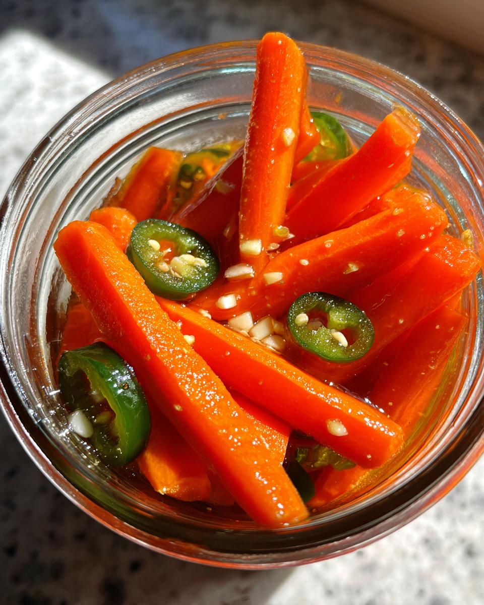 Close-up overhead view of bright orange pickled carrots sticks in a jar with sliced green jalapeños and minced garlic.