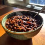 A steaming bowl of rich, dark black bean chili served in a green ceramic bowl on a wooden table.