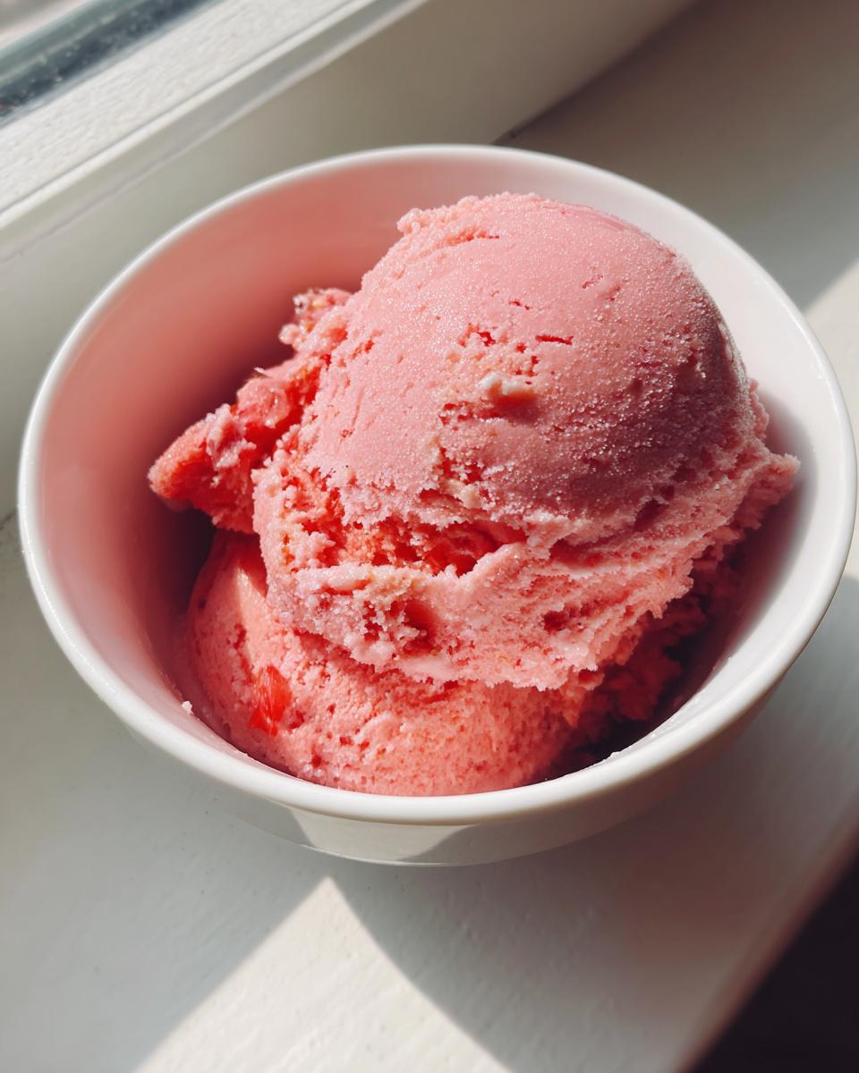 Two generous scoops of bright pink strawberry ice cream served in a white bowl, catching sunlight by a window.
